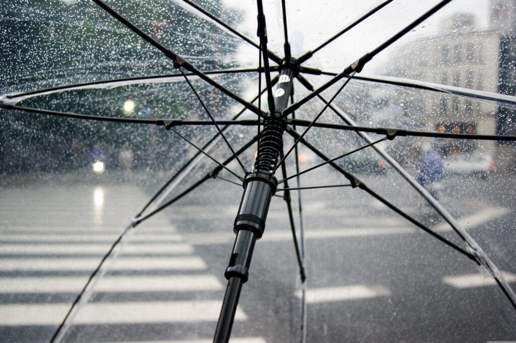 Close-up view from under a transparent umbrella with raindrops, looking out at a blurred city crosswalk and street in the background.