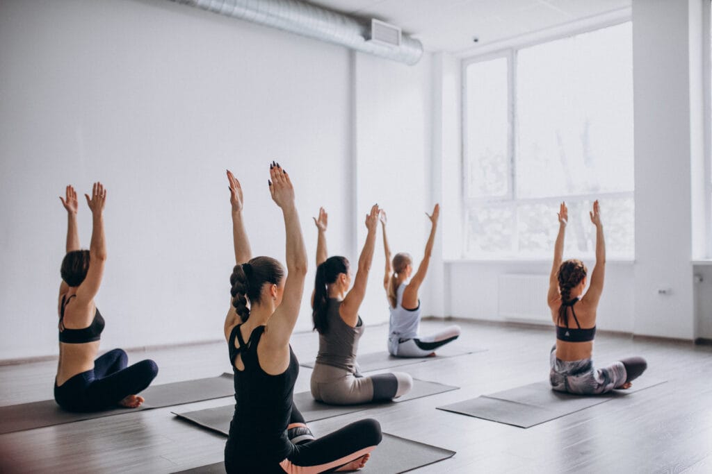 Five people sit on yoga mats in a bright studio, facing forward with arms raised overhead, participating in a group yoga class.