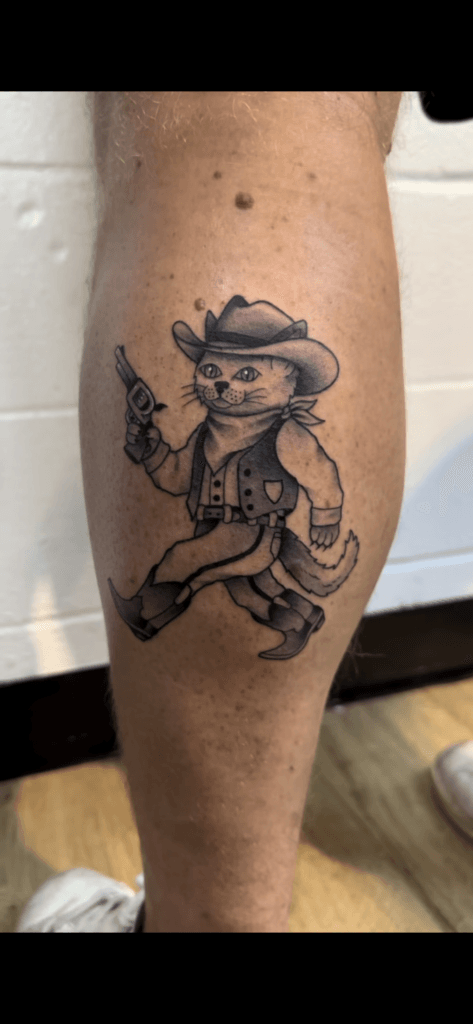 Cat dressed as a cowboy holding a gun, wearing a hat, vest, and bandana.