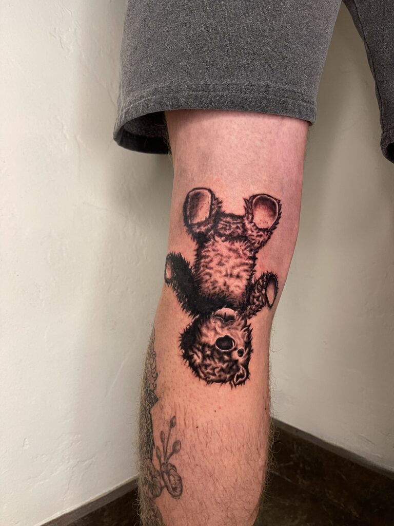 Detailed black and gray tattoo of a playful bear cub hanging upside down on a persons leg.