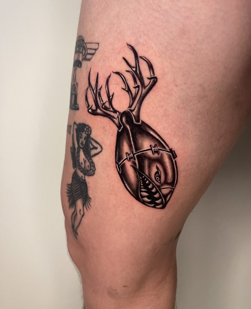 Detailed black and gray tattoo of a heart with antlers and a shark face on the thigh.