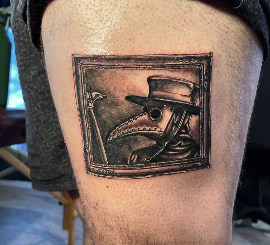 A detailed black and gray tattoo of a plague doctor mask with a wide-brimmed hat inside a decorative rectangular frame.