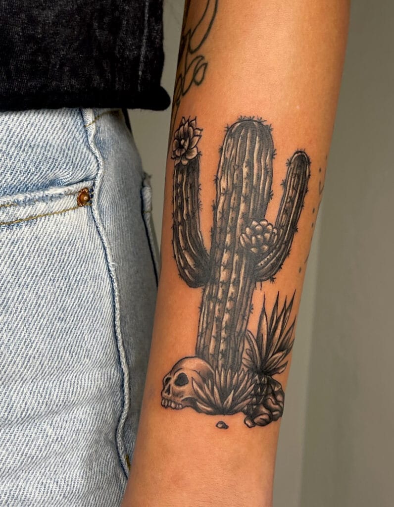 Black and gray cactus tattoo with a skull and desert plants on persons forearm.