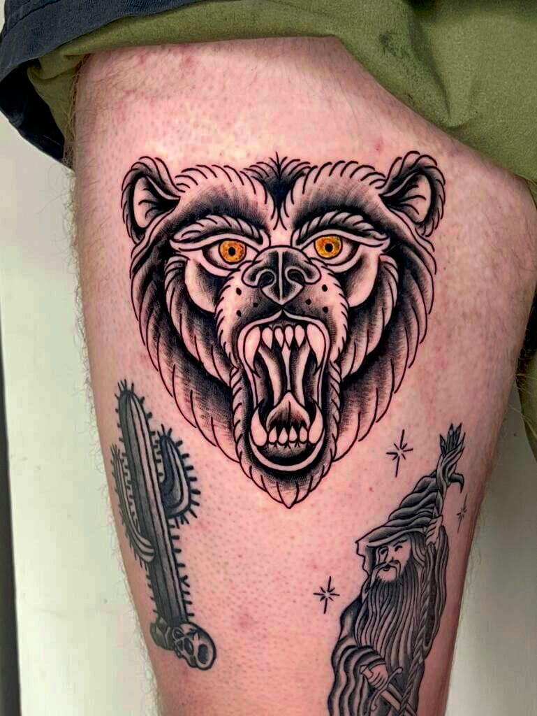 A fierce bear head tattoo with yellow eyes, an open mouth, and sharp teeth.