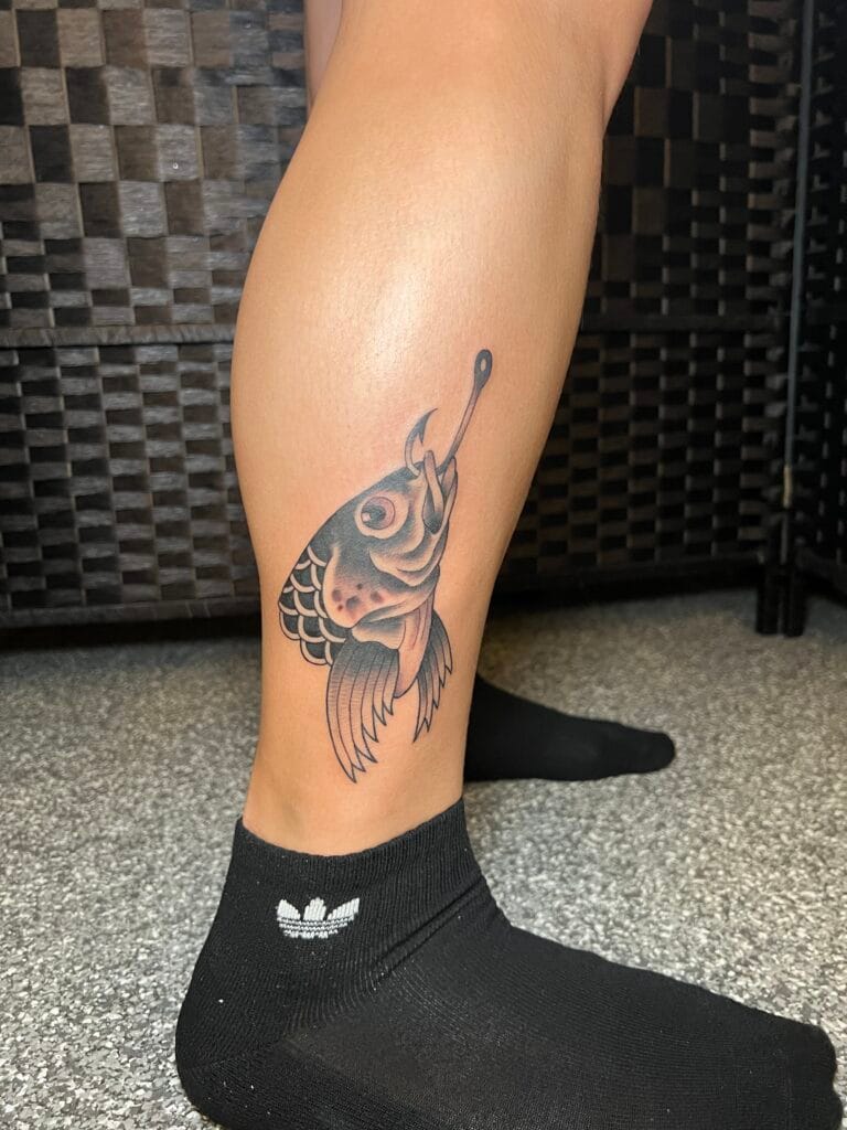 Tattoo of a fish with a fishing hook in its mouth on a persons lower leg.