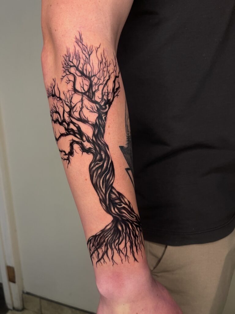 Black and gray tree tattoo with twisted trunk and sprawling branches on persons forearm.