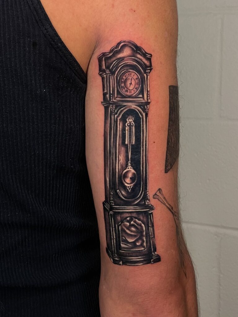Black and gray tattoo of a grandfather clock on persons upper arm, showing the clock face, pendulum, and base.