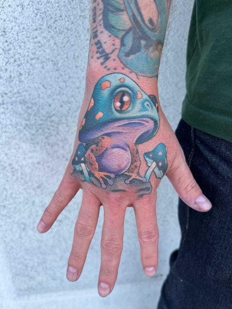 Colorful tattoo of a frog with large eyes on the hand, with part of an arm tattoo visible.