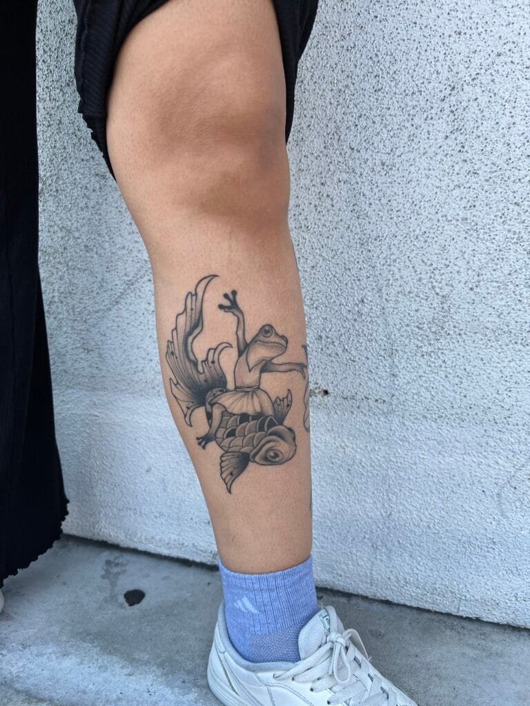 Tattoo of a frog with wings sitting on a fish, located on the lower leg.