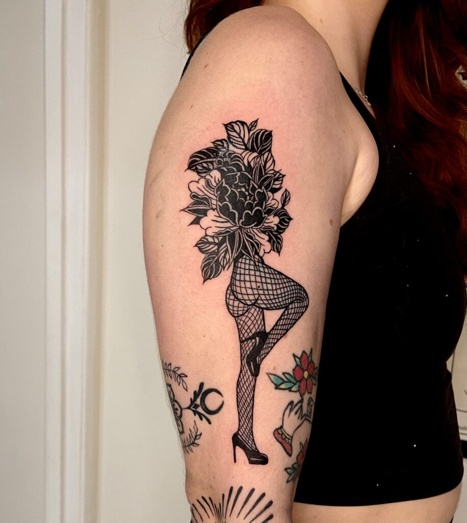 Tattoo of a woman with floral hair and fishnet stockings on upper arm.