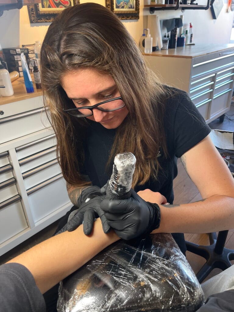Tattoo artist wearing black gloves and glasses, tattooing a persons forearm in a studio.