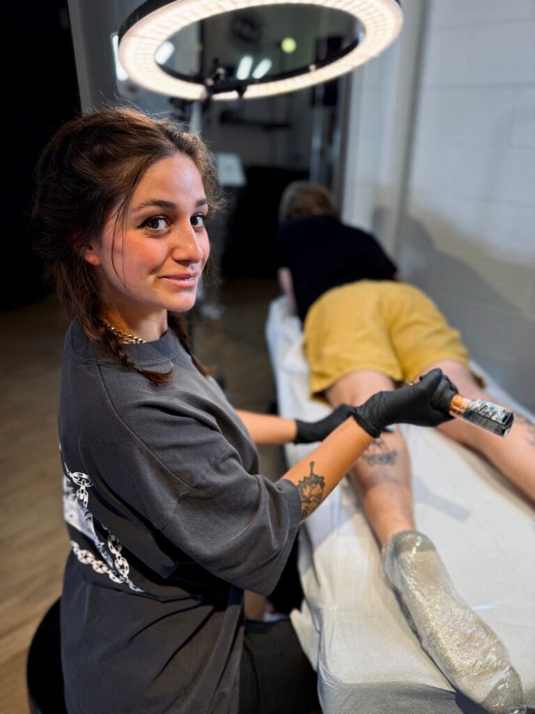 Tattoo artist with dark hair and black gloves applying ink to a persons leg in a tattoo studio.